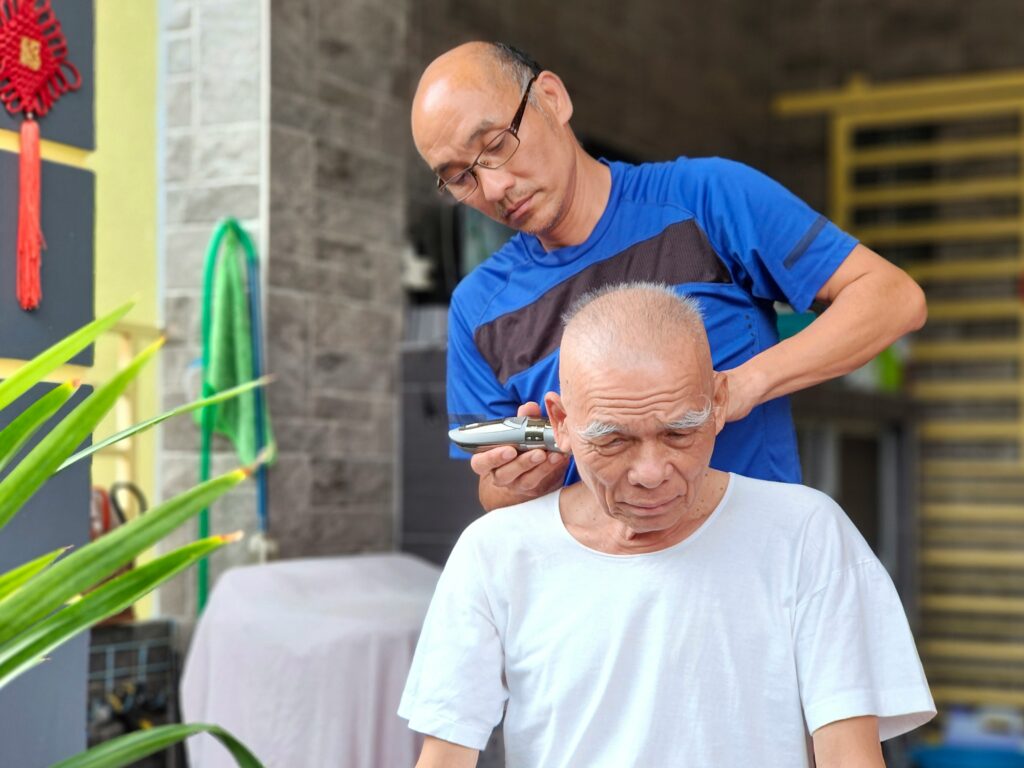 Son giving a haircut to his elderly father at home with electric clippers, showing family care and grooming.