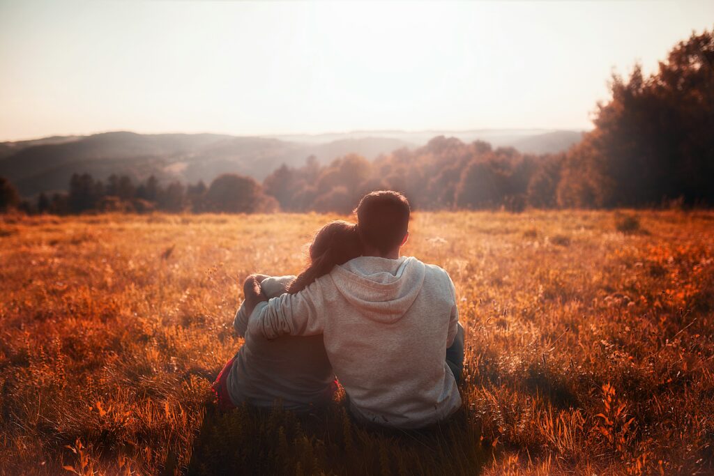 Romantic couple sitting in a field at sunset, enjoying a scenic mountain view and peaceful nature.
