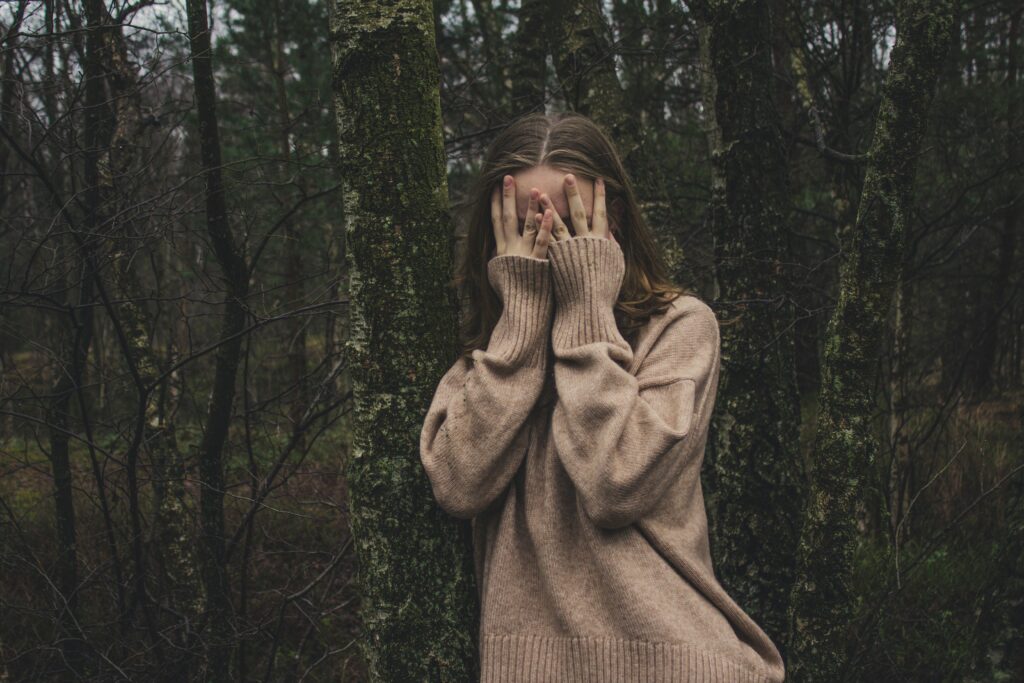 Young woman in a beige sweater covering her face with hands while standing against a tree in a forest, symbolizing anxiety, stress, or shyness in nature