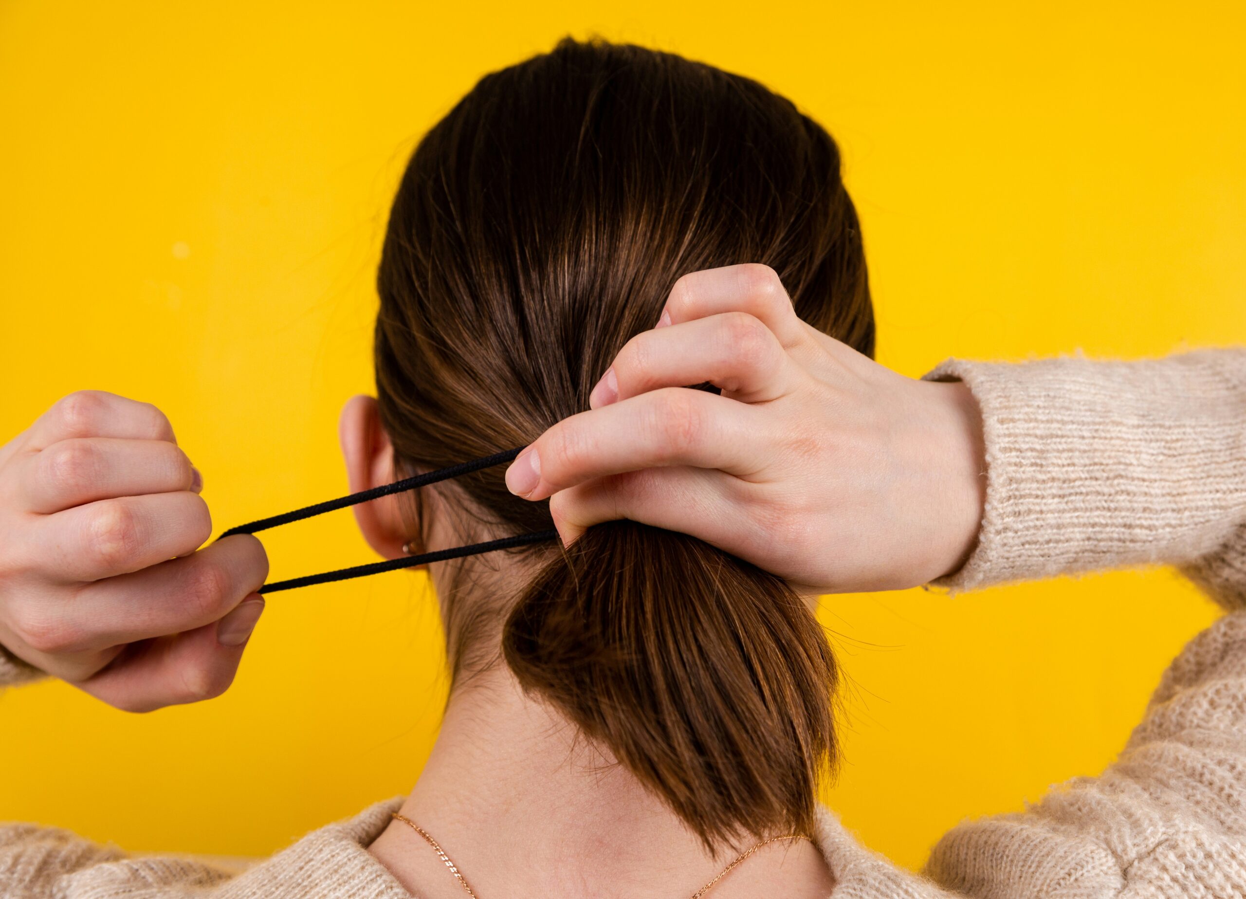 Close-up of a woman tying her brown hair into a ponytail with a black hair tie against a yellow background