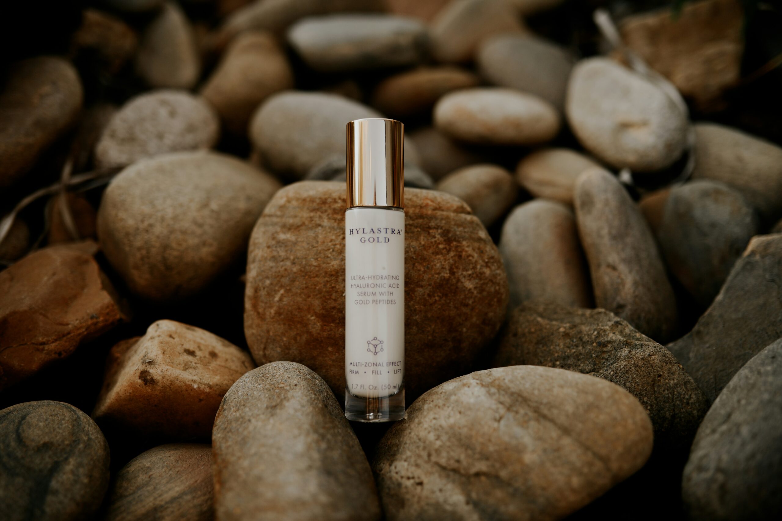 Hyalastra Gold ultra-hydrating hyaluronic acid serum with gold peptides displayed on natural river stones for beauty skincare product promotion.