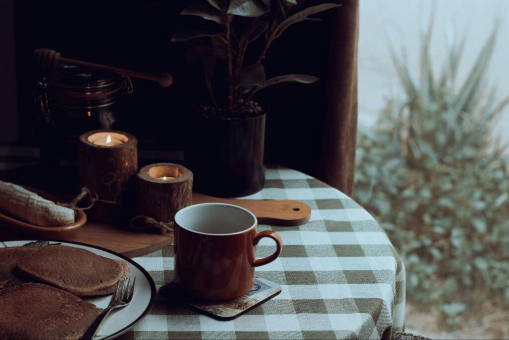 Cozy rustic breakfast with pancakes and coffee on a wooden table, candles lit beside a window with soft natural light.