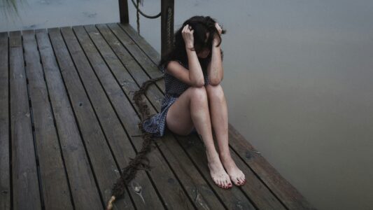 Sad woman sitting alone on a wooden dock by the water with her head in her hands, expressing loneliness, stress, or depression.